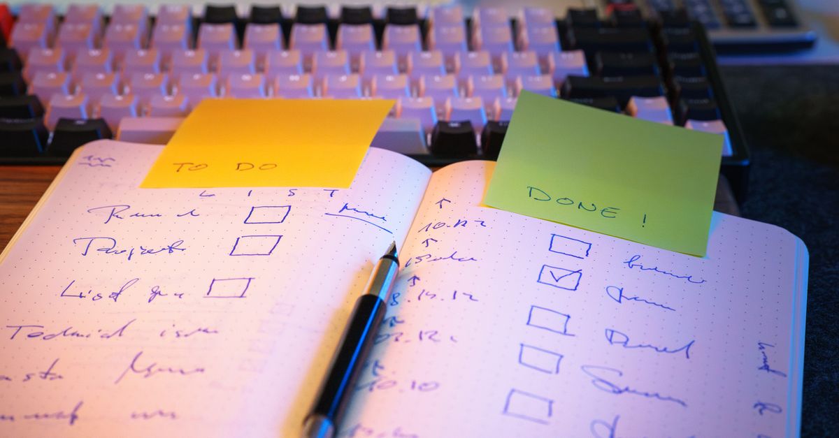 Open notebook with handwritten to-do and done checklists, sticky notes and a pen in front of a keyboard