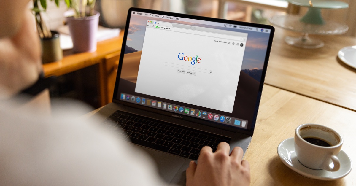 Person using a laptop open to the Google search home page, with a cup of coffee on a wooden table