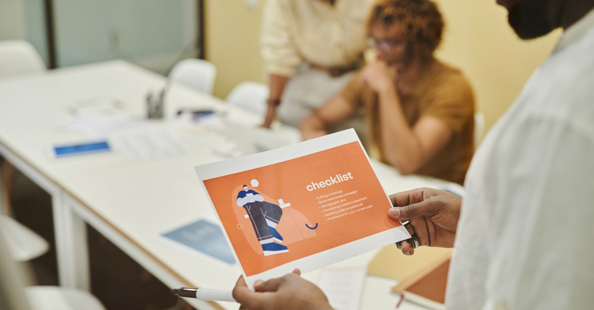 Man holds a printed checklist sheet while two colleagues sit at a white meeting table behind him