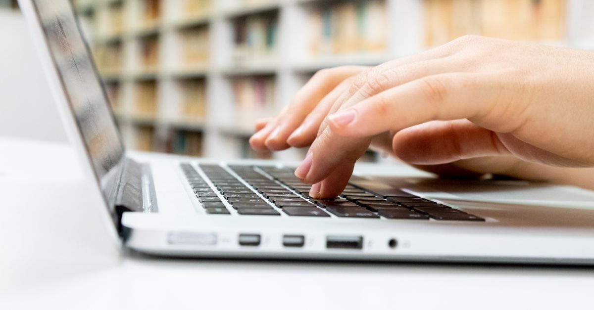 Hands typing on a silver laptop keyboard at a white desk with library bookshelves behind