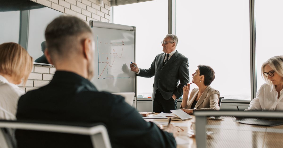 Senior executive in a suit presenting a chart on a flip chart to colleagues at a meeting table