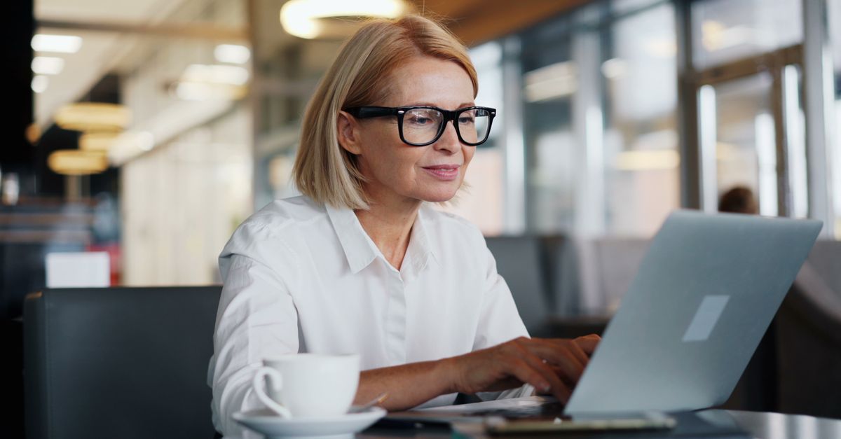Woman with glasses and a white shirt typing on a laptop at a cafe table beside a cup of coffee