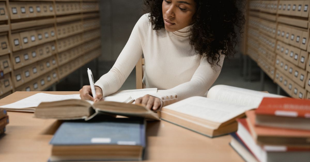 Young woman taking notes at a library desk surrounded by open books and archive drawers