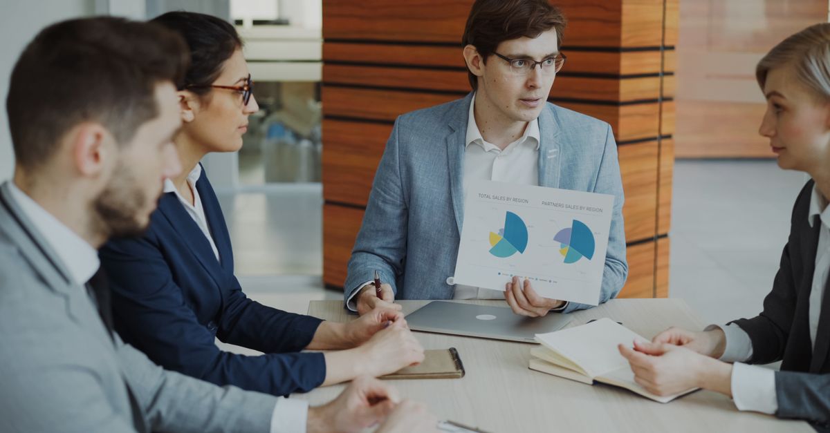 Man in a grey blazer shows a printed report with pie charts to three colleagues at a meeting table