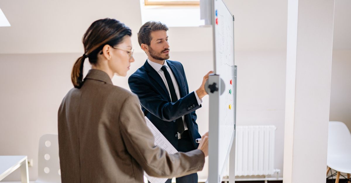 Two colleagues in business clothes discuss a plan at a whiteboard in a bright attic office