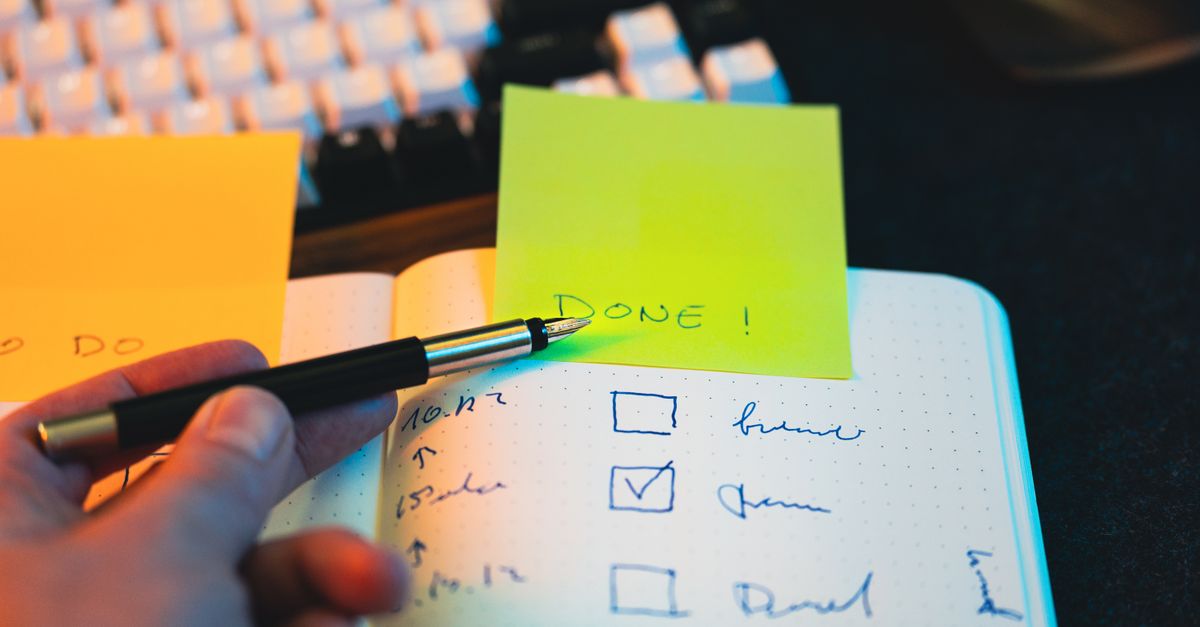 Hand holding a pen over a notebook checklist with one box ticked beside a sticky note that says done