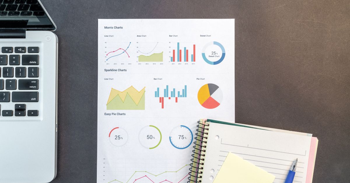 Printed sheet of line, bar and pie charts beside a laptop keyboard, notebook and pen on a dark desk