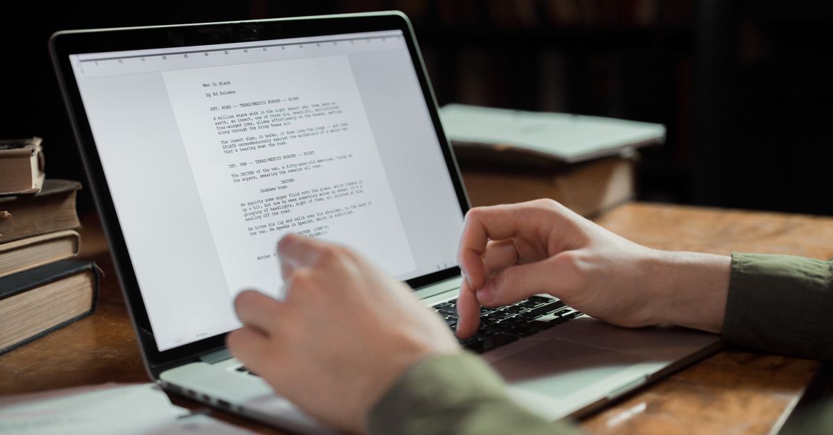 Hands typing a text document on a laptop at a wooden desk stacked with old books