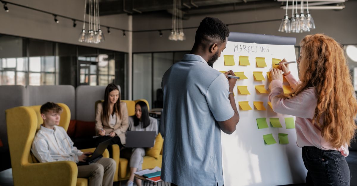 Two colleagues arranging sticky notes on a whiteboard while teammates work on sofas in an office