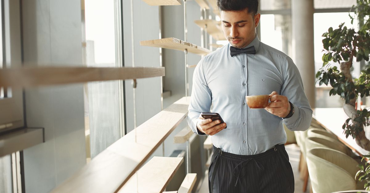 Man in a light blue shirt and bow tie reads his smartphone while holding a coffee cup in a bright cafe
