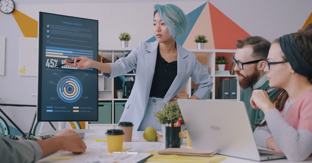 Woman presents a data dashboard on a large screen to two colleagues sitting at a table with a laptop